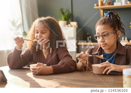 Portrait of two young African American girls eating cereal for breakfast at home lit by sunlight Portrait of two young African American girls eating cereal for breakfast at home lit by sunlight 114536103