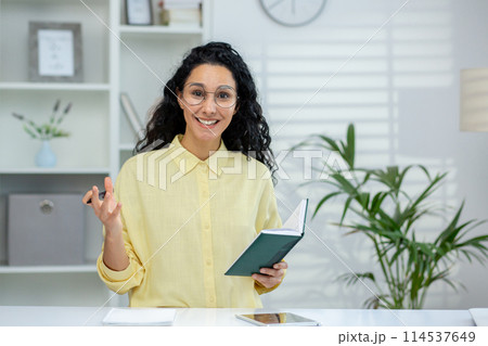 A smiling Hispanic woman conducting an online tutorial with a student, using gestures and holding a notebook in a bright office setting. 114537649