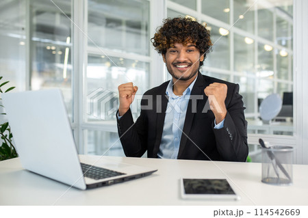 Cheerful businessman celebrating success at his desk in a modern office environment, expressing excitement and positive energy. Cheerful businessman celebrating success at his desk in a modern office environment, expressing excitement and positive energy. 114542669