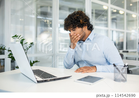 Young man feeling nausea while working at his desk in a modern office, indicating discomfort or illness, possibly related to stress or fatigue. Young man feeling nausea while working at his desk in a modern office, indicating discomfort or illness, possibly related to stress or fatigue. 114542690