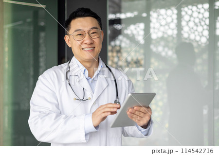 Asian mature male doctor in white coat using a tablet and smiling in a modern hospital or clinic setting, showcasing professional healthcare services. Asian mature male doctor in white coat using a tablet and smiling in a modern hospital or clinic setting, showcasing professional healthcare services. 114542716