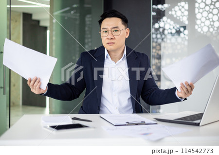 Asian mature man in a modern office looking frustrated while holding paperwork and documents, showing stress and confusion. 114542773