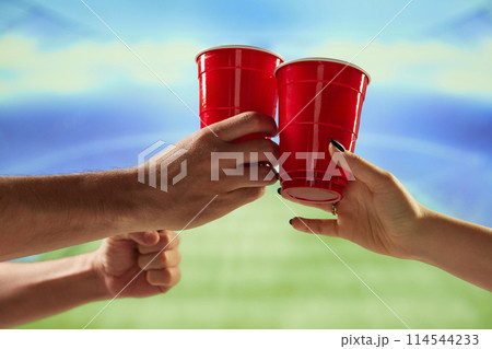 Hands holding red mugs with beer and clicking with blurred sport open air stadium on background 114544233
