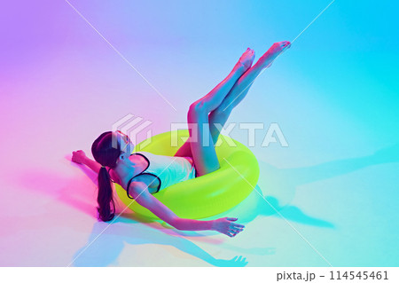 Woman in retro bikini enjoys her travelling like swimming in beach in yellow float in neon light against vibrant gradient background. 114545461