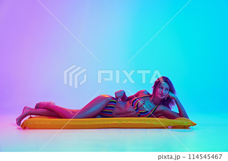 Young tender woman lying on air mattress and relaxing like swimming in ocean in neon light against vibrant gradient background. Young tender woman lying on air mattress and relaxing like swimming in ocean in neon light against vibrant gradient background. 114545467