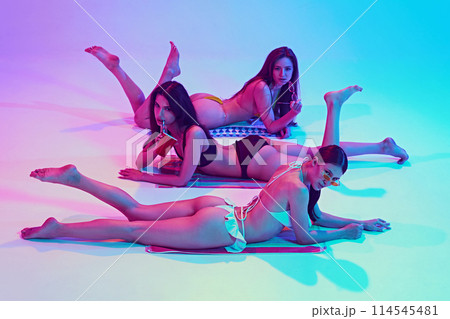 Three young, fit women lounge in style swimwear in neon light against vibrant gradient background. Summer chic. Three young, fit women lounge in style swimwear in neon light against vibrant gradient background. Summer chic. 114545481