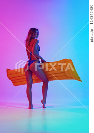 Rear view photo of young woman in swimsuit holds inflatable in neon light against vibrant gradient background. Summer vibe. Rear view photo of young woman in swimsuit holds inflatable in neon light against vibrant gradient background. Summer vibe. 114545486