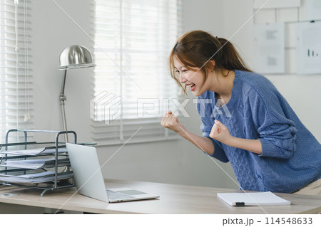 Triumphant Asian businesswoman celebrating professional victory with arms raised in front of a laptop at home office. Triumphant Asian businesswoman celebrating professional victory with arms raised in front of a laptop at home office. 114548633