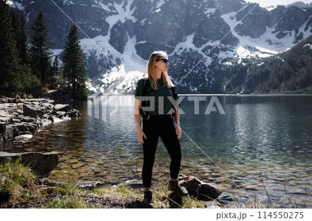 Blonde woman with backpack standing at full height by lake, mountains and snow in backdrop Blonde woman with backpack standing at full height by lake, mountains and snow in backdrop 114550275