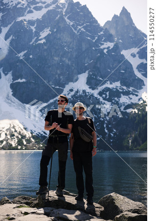 Two men on rock by lake, mountains in background, enjoying natural landscape Two men on rock by lake, mountains in background, enjoying natural landscape 114550277