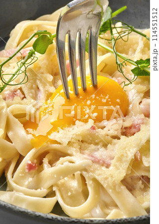 Fork breaking golden egg yolk on carbonara pasta 114551512