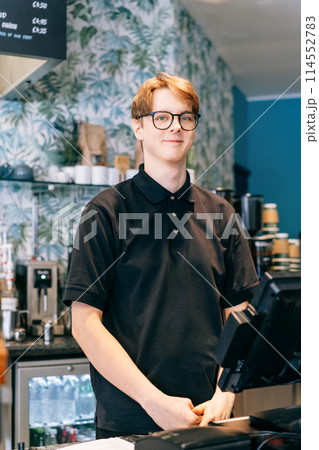 Young waiter serving customer at cash point in cafe. Man working with POS terminal. Cashier, barista checking for payment receipt. Hospitality, server and preparing a slip at the till in coffee shop. 114552783