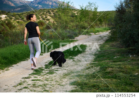 Full size shot of a happy woman smiling looking away, walking her dog in mountains nature outdoors. Rear view Full size shot of a happy woman smiling looking away, walking her dog in mountains nature outdoors. Rear view 114553254