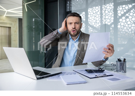 Shocked businessman holding a document while sitting at his desk in an office. He appears surprised and worried, surrounded by paperwork and a laptop. Shocked businessman holding a document while sitting at his desk in an office. He appears surprised and worried, surrounded by paperwork and a laptop. 114553634