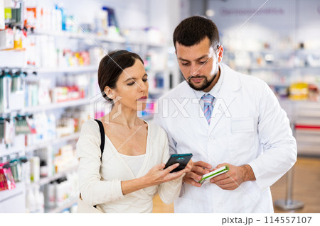 Female shopper shows name of drug on smartphone screen. Pharmacist helps to find medicine in pharmacy 114557107