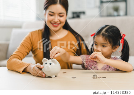 Cute little asian girl saving money by adding a coin in piggy bank with mother at home 114558380