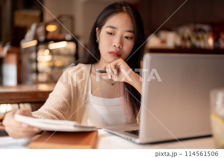 Young focused Asian woman working remotely at a coffee shop, taking notes in her notebook. 114561506