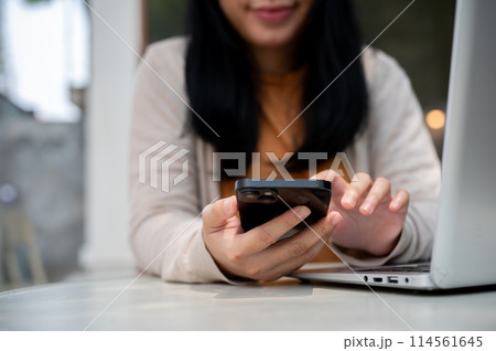 An Asian woman checking messages on her smartphone while working on her laptop at a cafe. An Asian woman checking messages on her smartphone while working on her laptop at a cafe. 114561645