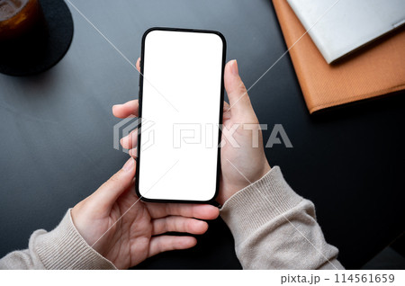 Top view image of a woman holding a smartphone white-screen mockup over an office desk workspace. 114561659