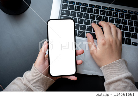 A top view image of a woman typing on laptop keyboard and holding a white-screen smartphone mockup. 114561661