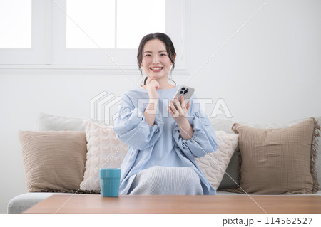 綺麗なリビングでスマホを持って考える若い女性　画面を見て笑顔で何かを見つける　正面　カメラ目線 114562527