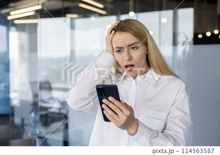 Stressed young businesswoman in a modern office, reacting to a surprising text message on her smartphone with a concerned expression. 114563587