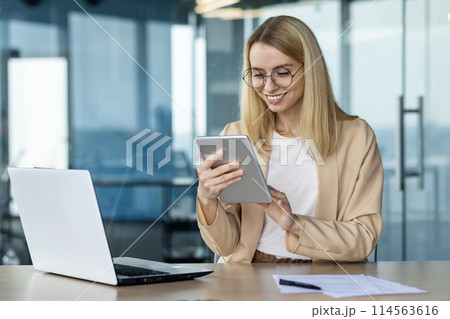 A smiling professional woman in a beige blazer uses a tablet while sitting at a glass table in a contemporary office setting, with a laptop and documents. 114563616