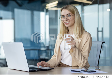 A professional young businesswoman enjoys a moment of hydration while working on her laptop in a brightly lit modern office. 114563618