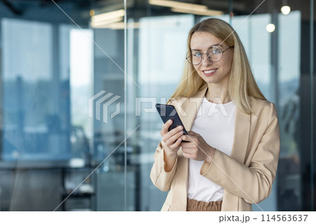 Confident young businesswoman using a smartphone in a modern office. She is smiling and wearing glasses, portraying a friendly and professional appearance. Confident young businesswoman using a smartphone in a modern office. She is smiling and wearing glasses, portraying a friendly and professional appearance. 114563637
