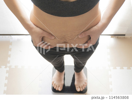 産後の妊娠線が残るお腹の肉をつまみながら体重計に乗る女性 114563808