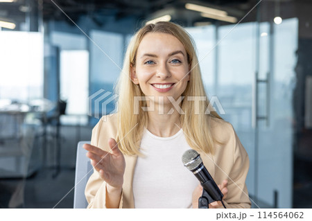 Smiling businesswoman holding a microphone during a presentation in a modern office. Professional woman delivering a speech or lecture. 114564062