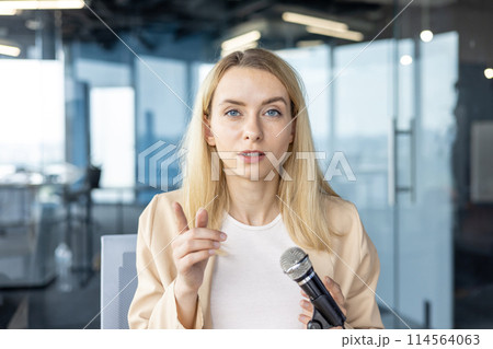 Professional female in a light blazer speaks into a microphone, engaging her audience in a contemporary office setting. Her expression is focused and communicative. 114564063