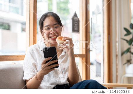 A young, happy Asian woman enjoying a donut while using her smartphone on a sofa in a cafe. A young, happy Asian woman enjoying a donut while using her smartphone on a sofa in a cafe. 114565588
