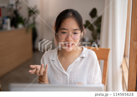 A young Asian woman is talking, having an online meeting while working remotely at a coffee shop. 114565628