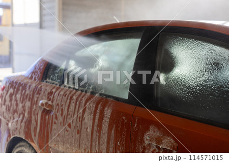 A driver washes off foam from an orange car at a car wash with water from a pressure gun. A powerful jet of water effectively removes dirt from the car body. A man takes care of his car. A driver washes off foam from an orange car at a car wash with water from a pressure gun. A powerful jet of water effectively removes dirt from the car body. A man takes care of his car. 114571015