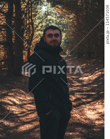 Full length portrait of a man in the forest. A brutal man looks thoughtfully at the camera while standing on a forest path surrounded by evergreen pines. Concept portrait of a man in the forest. 114571036
