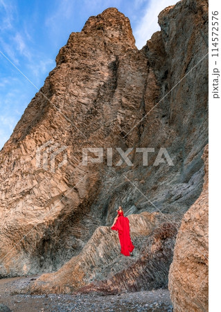 woman sea red dress. Woman with long hair on a sunny seashore in a red flowing dress, back view, silk fabric waving in the wind. Against the backdrop of the blue sky and mountains on the seashore. 114572576