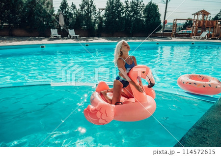 A woman is sitting on a pink flamingo float in a pool. The pool is surrounded by chairs and umbrellas. 114572615
