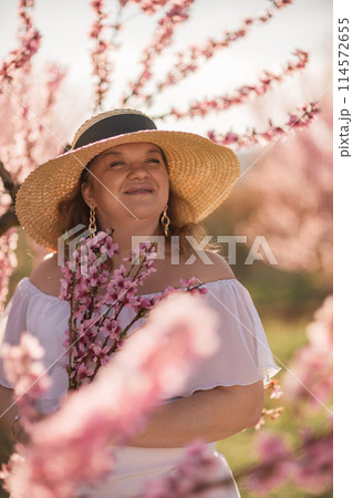 Woman blooming peach orchard. Against the backdrop of a picturesque peach orchard, a woman in a long white dress and hat enjoys a peaceful walk in the park, surrounded by the beauty of nature. 114572655