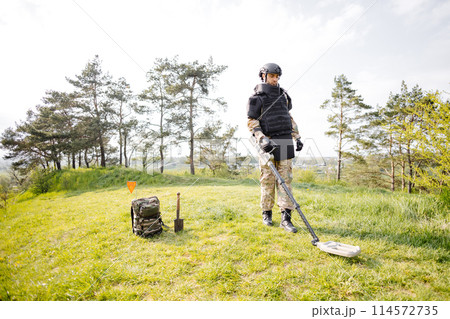 A man in a military uniform and bulletproof vest works in the forest with a metal detector. A minesweeper performs work on demining the territory 114572735