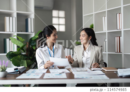Two young businesswomen discussing reports and smiling at the office Two young businesswomen discussing reports and smiling at the office 114574985