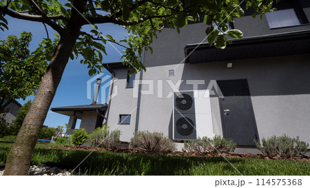 The outdoor unit of a heat pump is mounted against the wall of a modern cottage. The outdoor unit of a heat pump is mounted against the wall of a modern cottage. 114575368