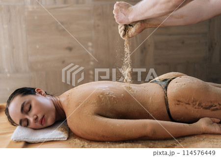 Professional spa technologist pouring heated sand on back of female client to scrubbing in luxury wellness center. Young woman having massage in spa salon. Concept of body care and rehabilitation. 114576449