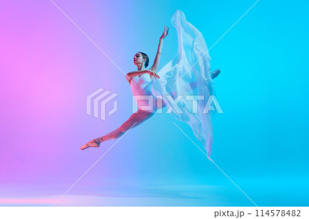 Ballet dancer performs jumping split in motion with flowing white cloth in neon light against vivid gradient background. Ballet dancer performs jumping split in motion with flowing white cloth in neon light against vivid gradient background. 114578482