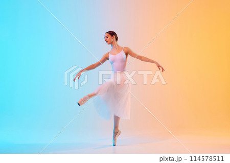 Young ballerina in white tutu an pointe practicing dance moves in neon light against blue-orange gradient background. Young ballerina in white tutu an pointe practicing dance moves in neon light against blue-orange gradient background. 114578511