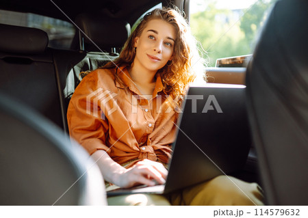 A young curly woman with a laptop sitting in back seat of a car. Beautiful woman uses laptop in car. A young curly woman with a laptop sitting in back seat of a car. Beautiful woman uses laptop in car. 114579632