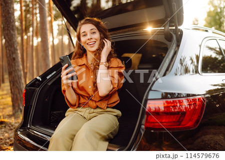 Young female tourist in casual clothes sits in the trunk of a car and uses a phone. Adventure. 114579676