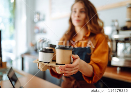 Cheerful woman in an apron at the bar counter holds coffee glasses in a cafe. Takeaway food concept. 114579886