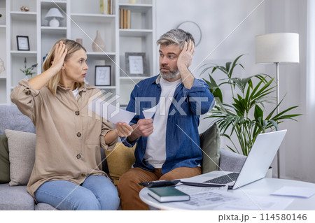 Stressed couple sitting on the couch discussing bills and finances. They appear worried while holding papers in their hands and looking at each other. 114580146