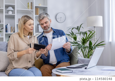 Mature couple sitting on a couch, reviewing documents and using a tablet at home. They seem focused and engaged in a financial discussion. 114580148